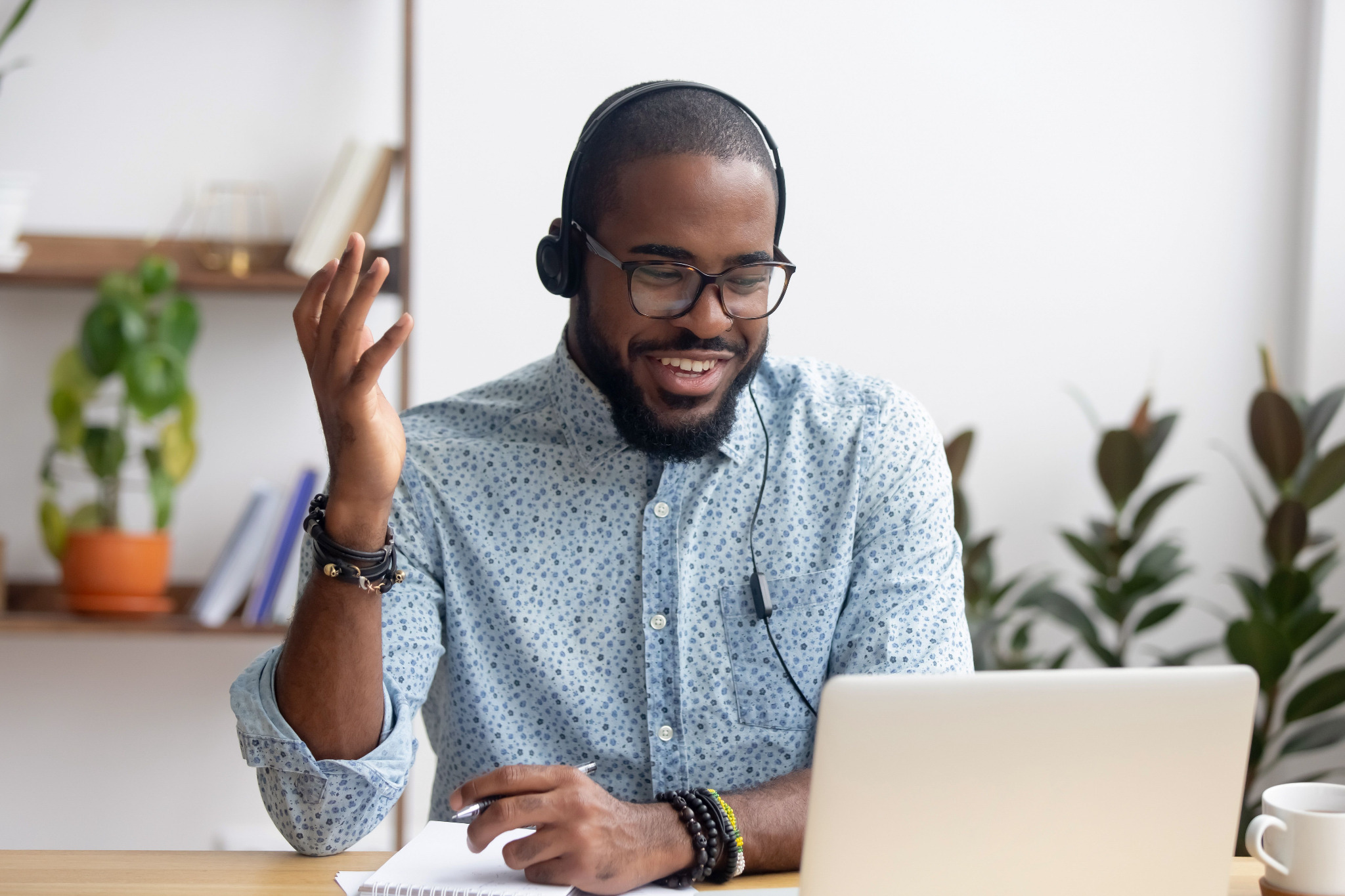 A cheerful man with a beard wearing a headset and glasses, gesturing while looking at a laptop, seated at a desk in a room with plants in the background.